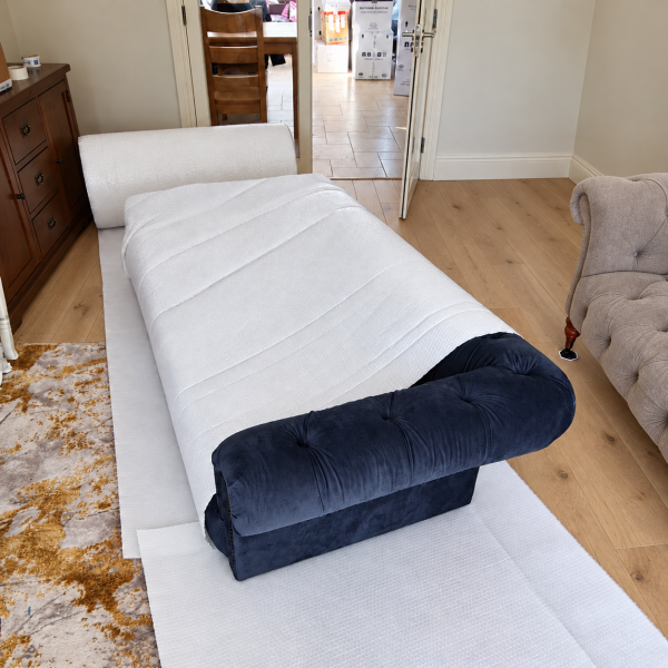 White export bubble blanket covering a navy sofa, with the near arm exposed to show the protective wrapping.